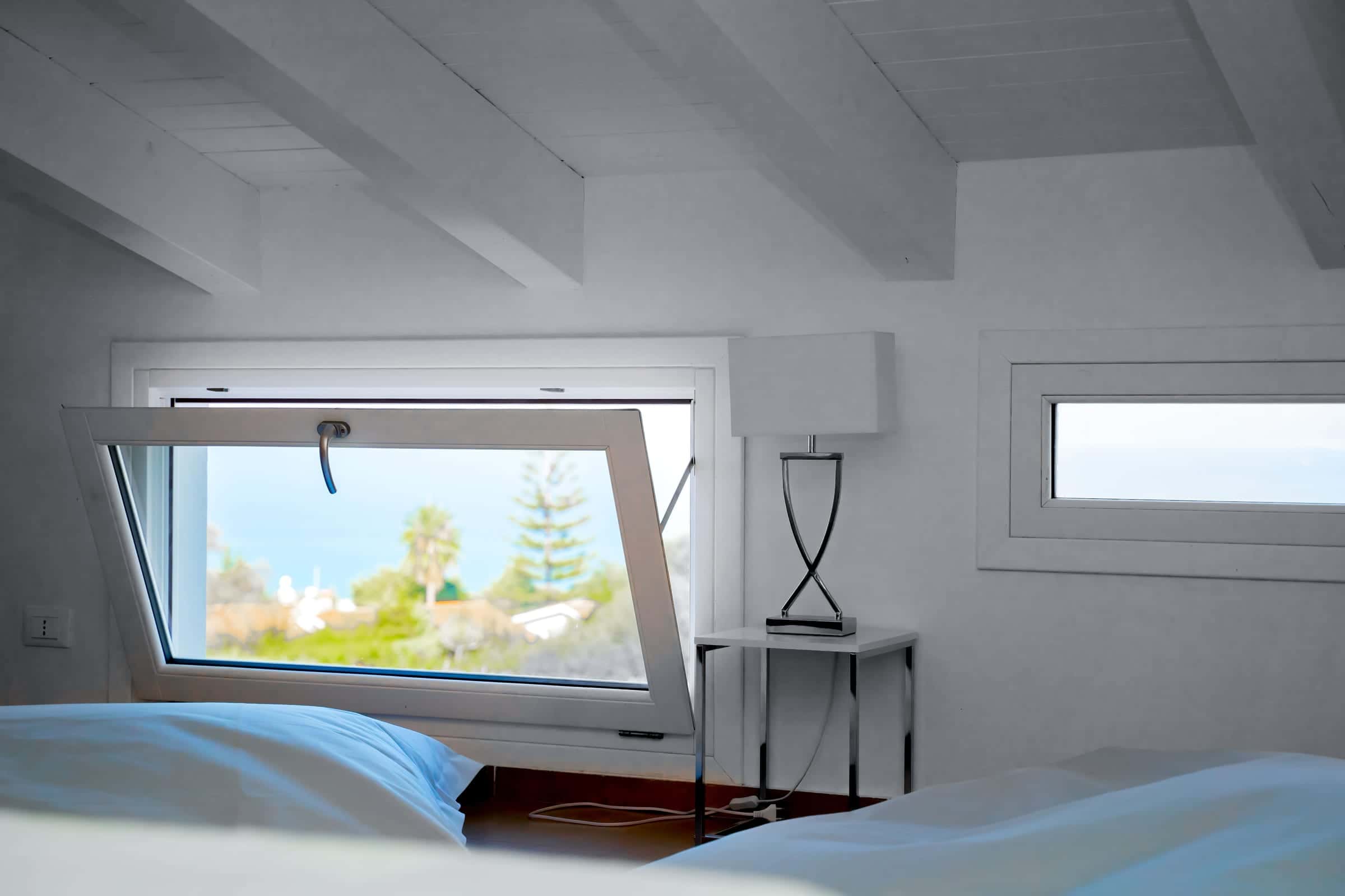 Bedroom under a white beamed ceiling; the tilted window looks out over trees to the sea