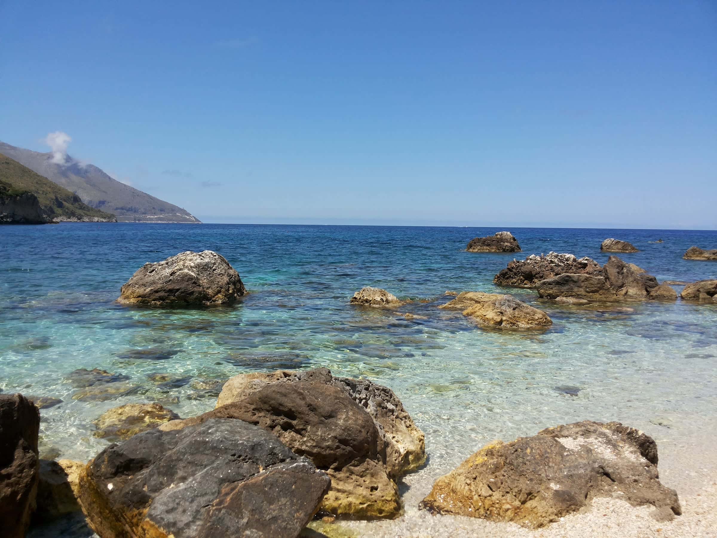 Clear water and boulders at Cala Mazzo di Sciacca near Scopello, with mountains along the coast in the distance