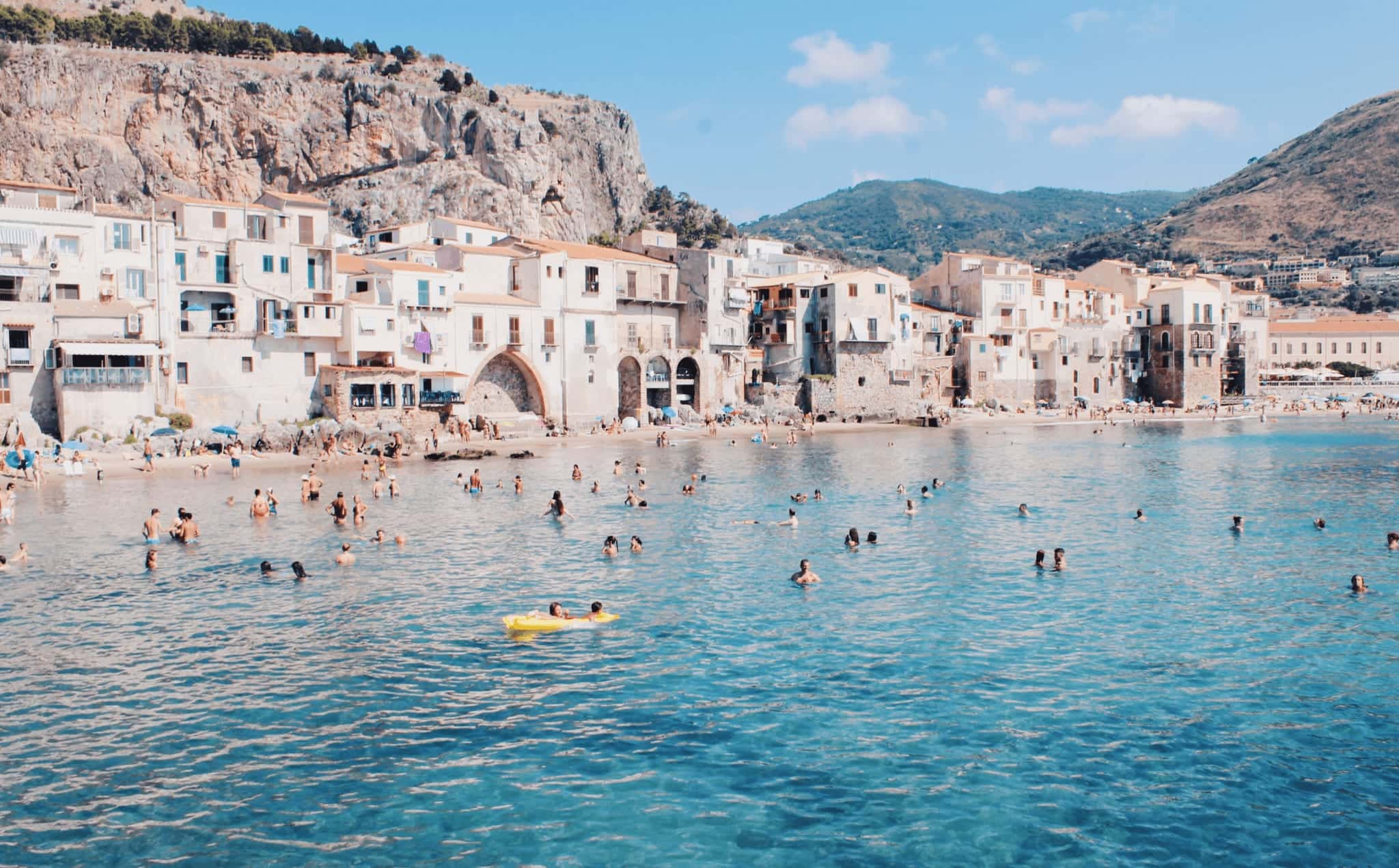Swimmers in clear water off Cefalù, with the old town on the shore and the rock above it