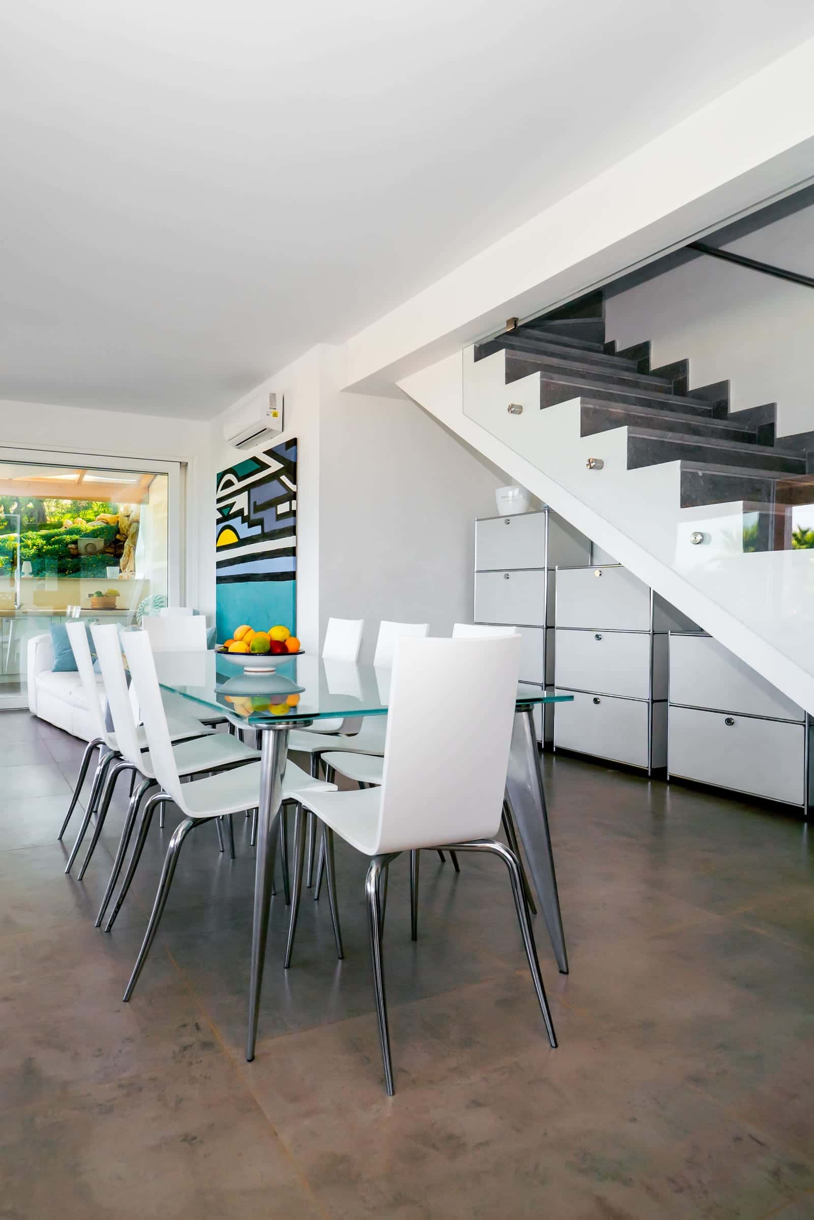 Dining area with a glass table and white chairs beside a staircase with a glass balustrade