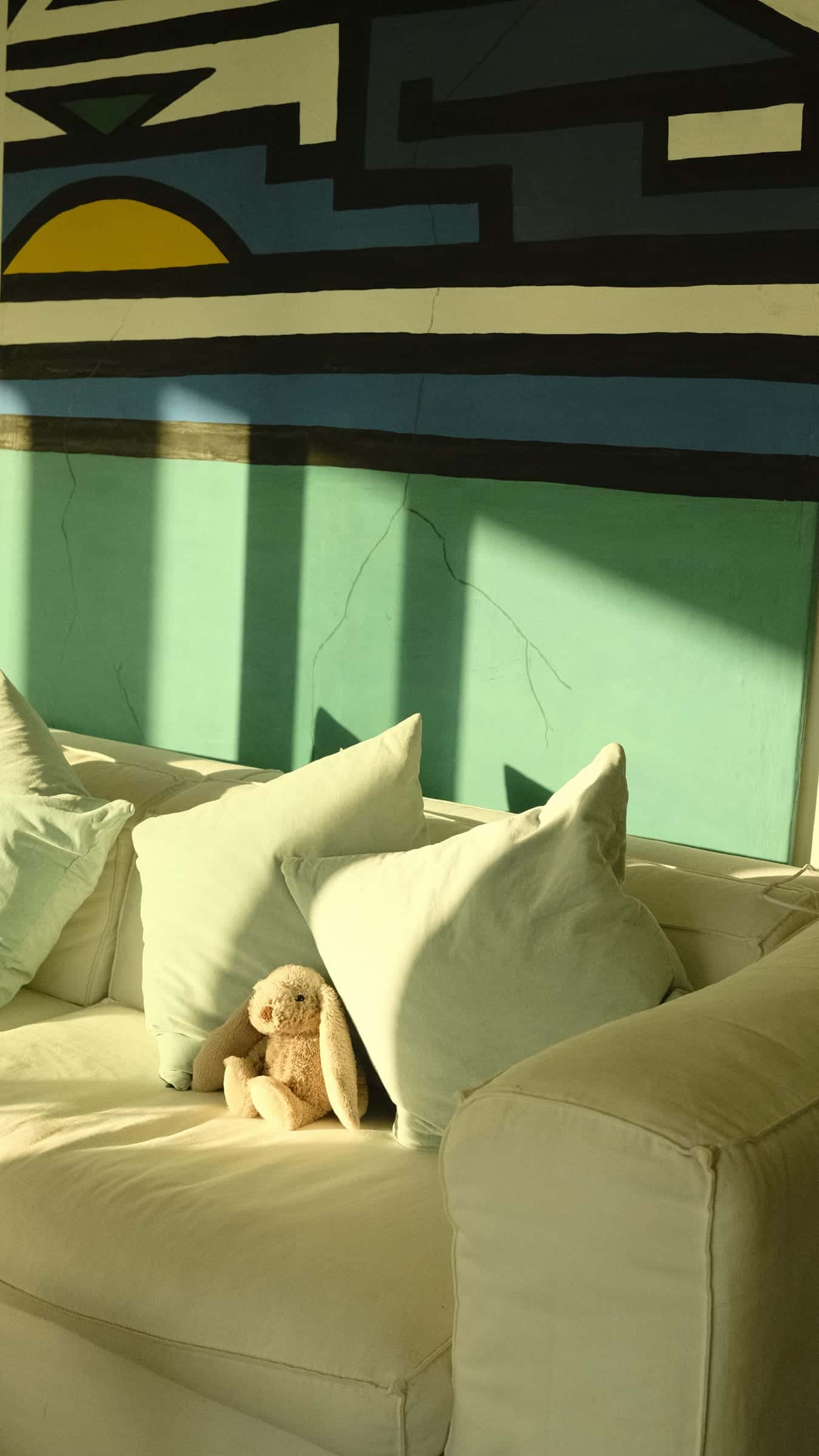 Cushions and a toy rabbit on the white sofa, below a large geometric wall painting in green, blue, black and yellow