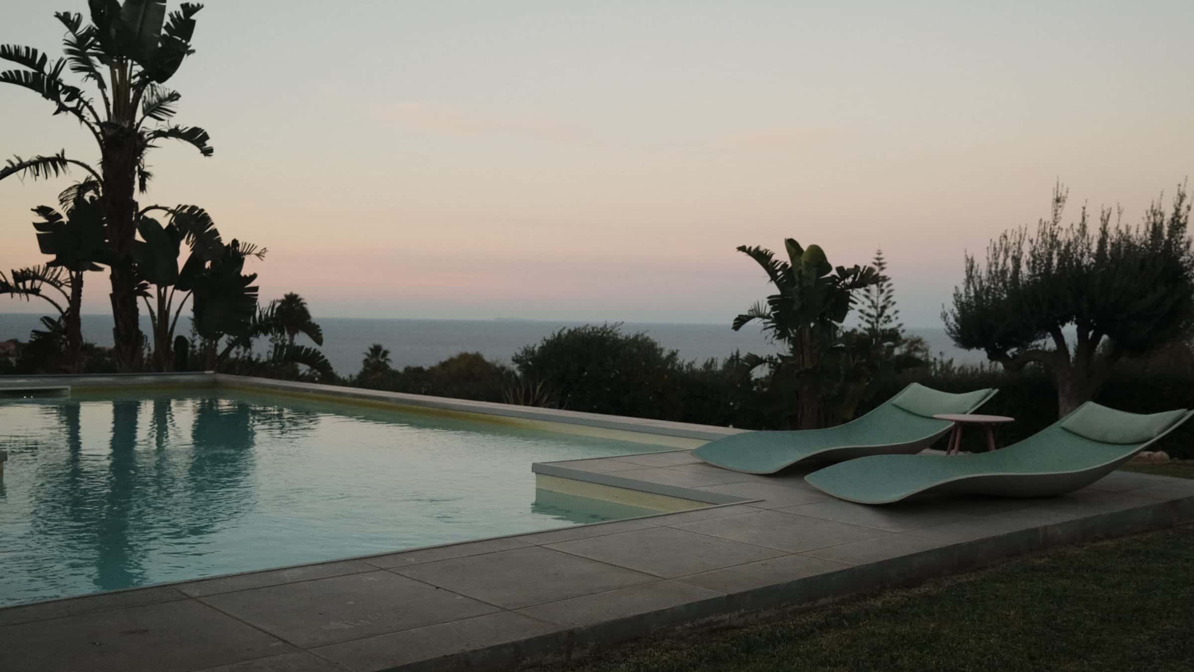 The pool at dusk with two sun loungers on its edge, tall tropical plants on the left and the sea under a pink sky