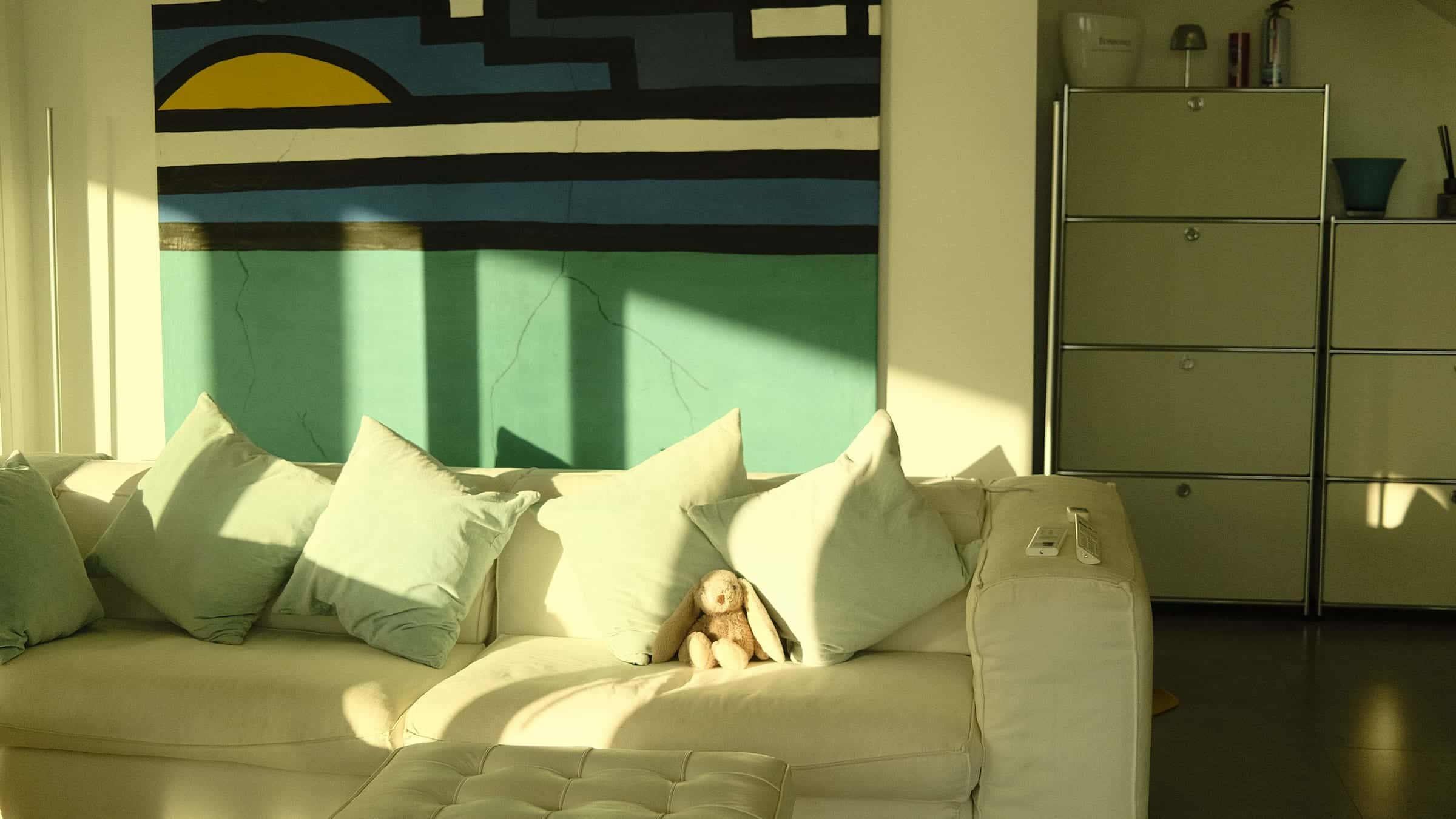 Living room in low sun: the white sofa below a large geometric wall painting, metal cabinets beside it on a dark tiled floor