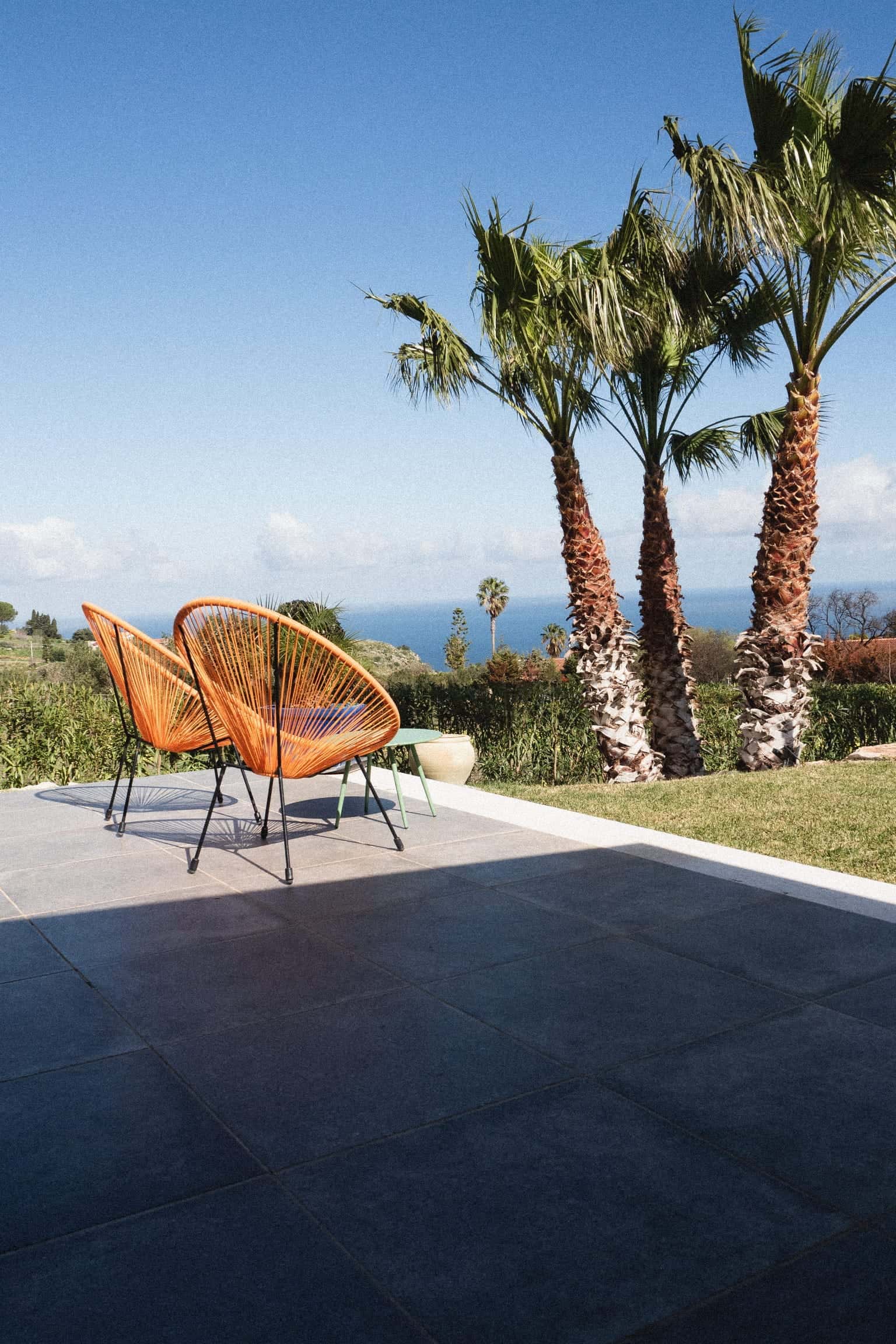 Two orange chairs with woven seats on a shaded terrace, three palms on the lawn beside it and the sea under a blue sky