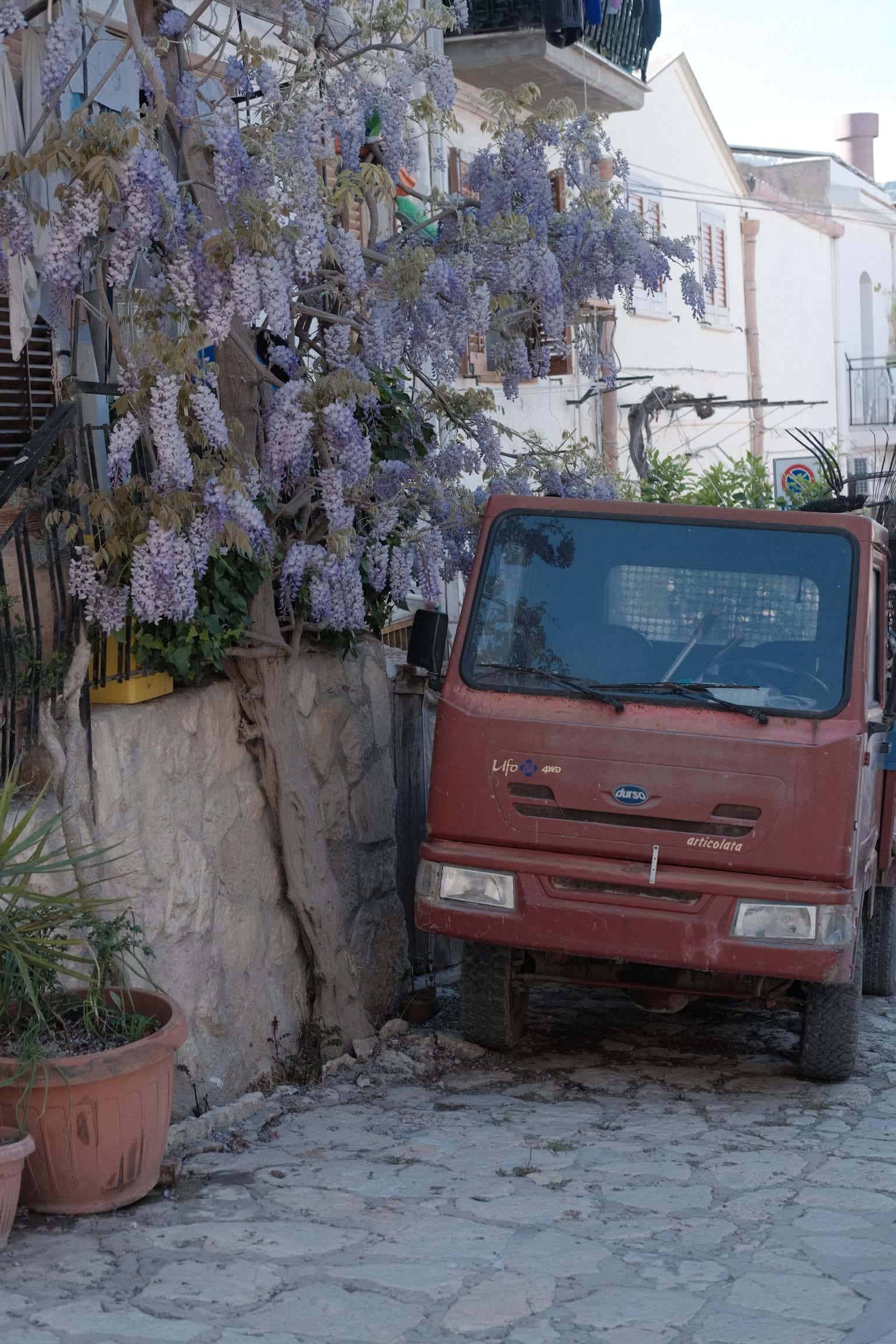 Purple wisteria over a stone wall in a cobbled lane, a small red truck parked beside it and white houses behind