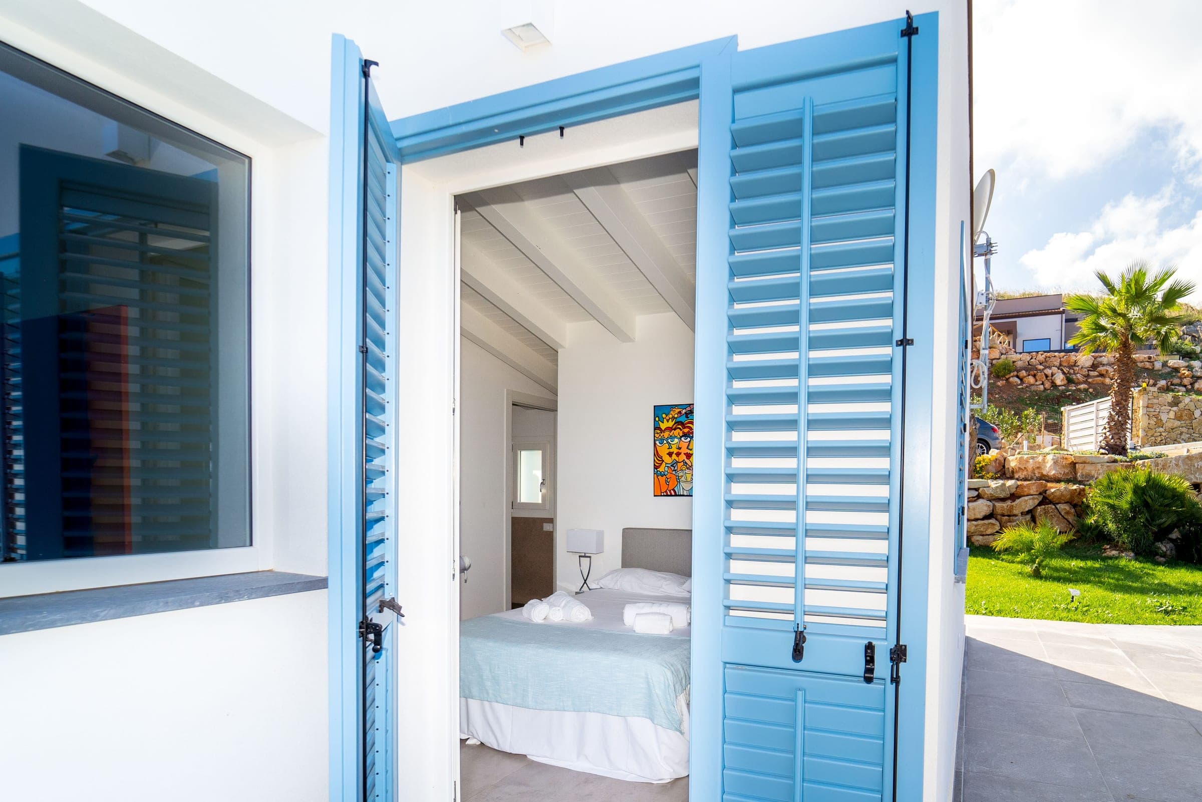 Open blue louvred shutters and, beyond them, a bedroom with a double bed under a white beamed ceiling