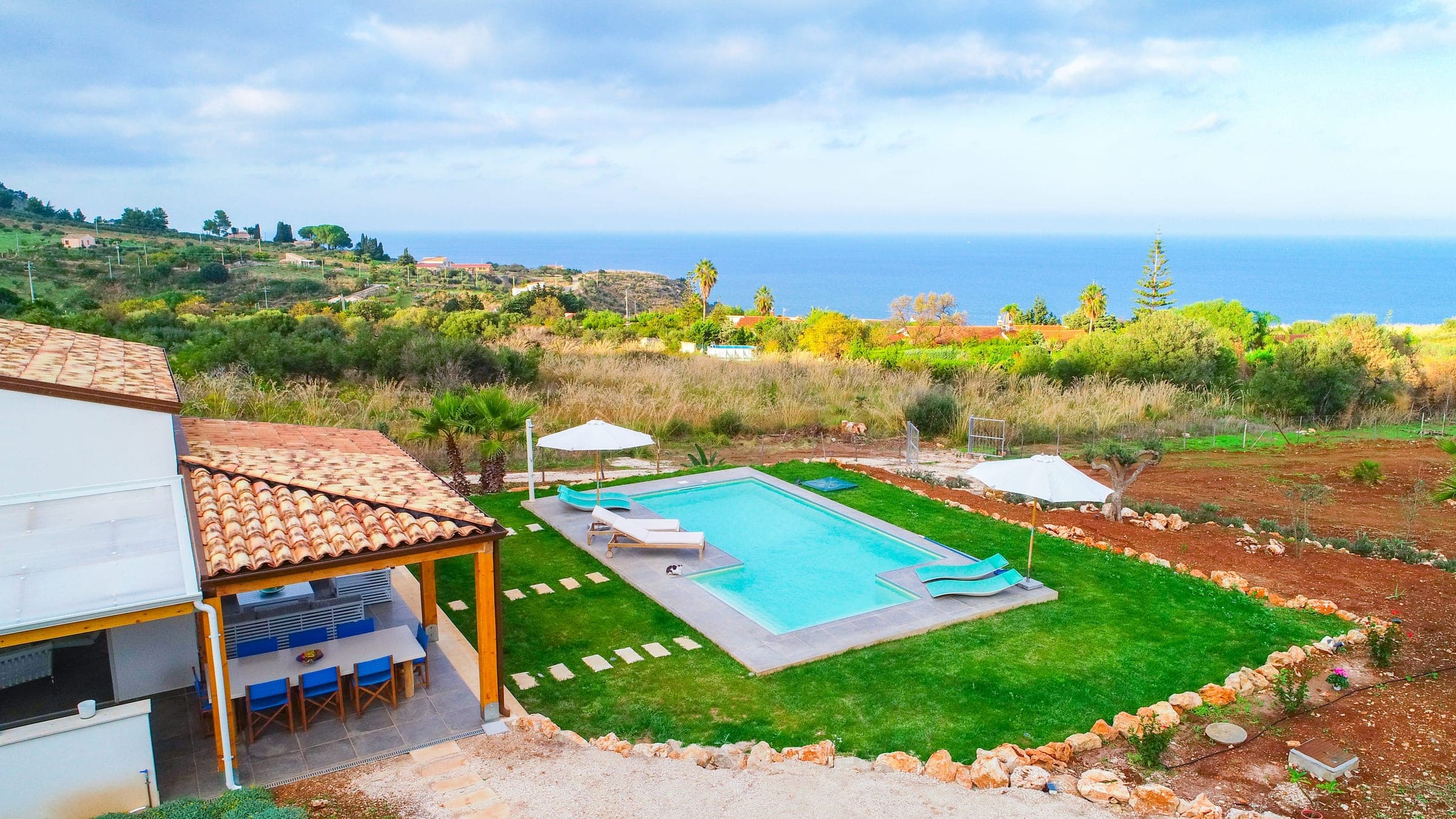 The pool and its sun loungers in the lawn, the covered dining terrace on the left and the sea on the horizon