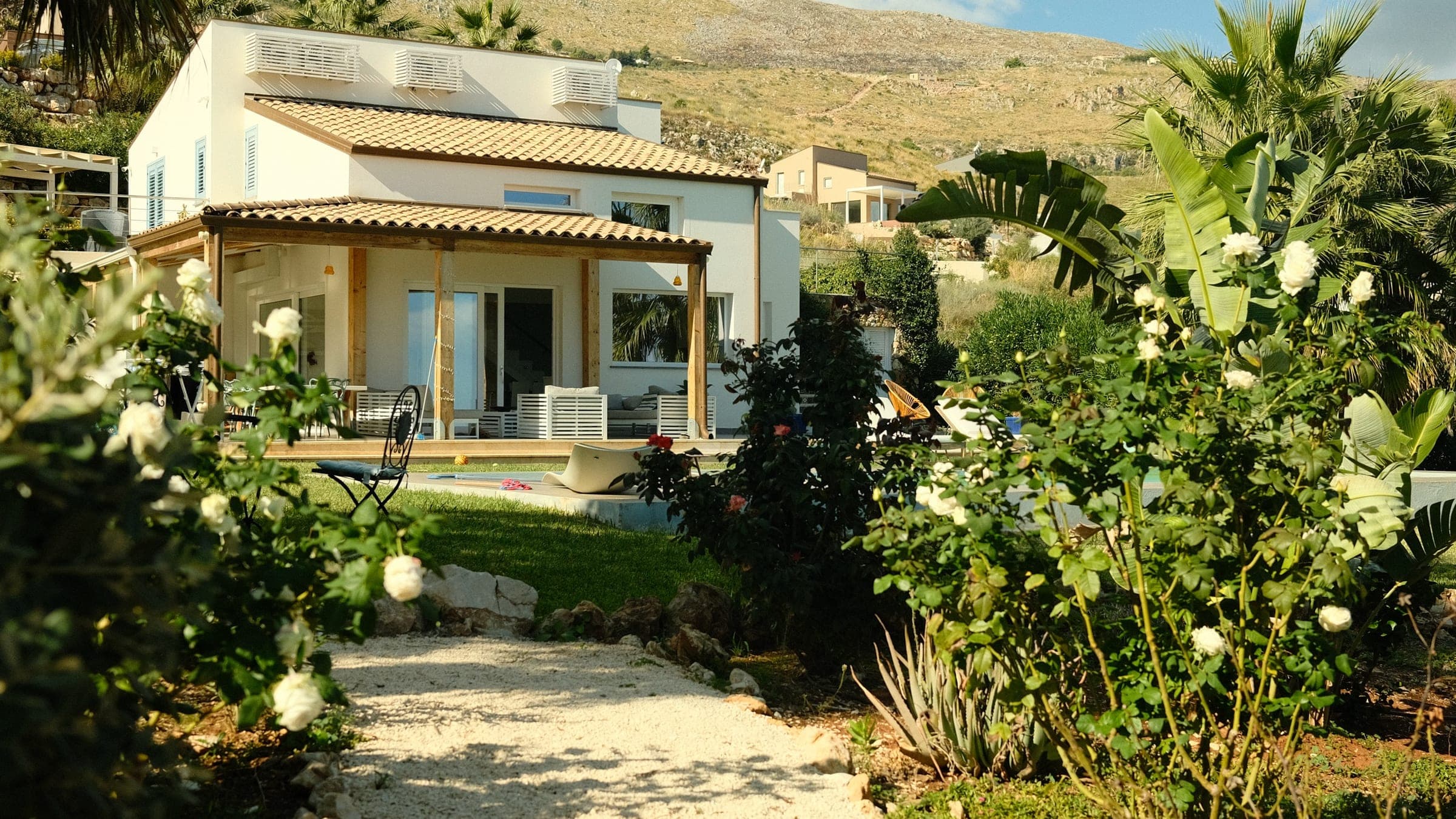 Gravel path between white roses and banana plants leading to the white villa with its terracotta roof and pergola