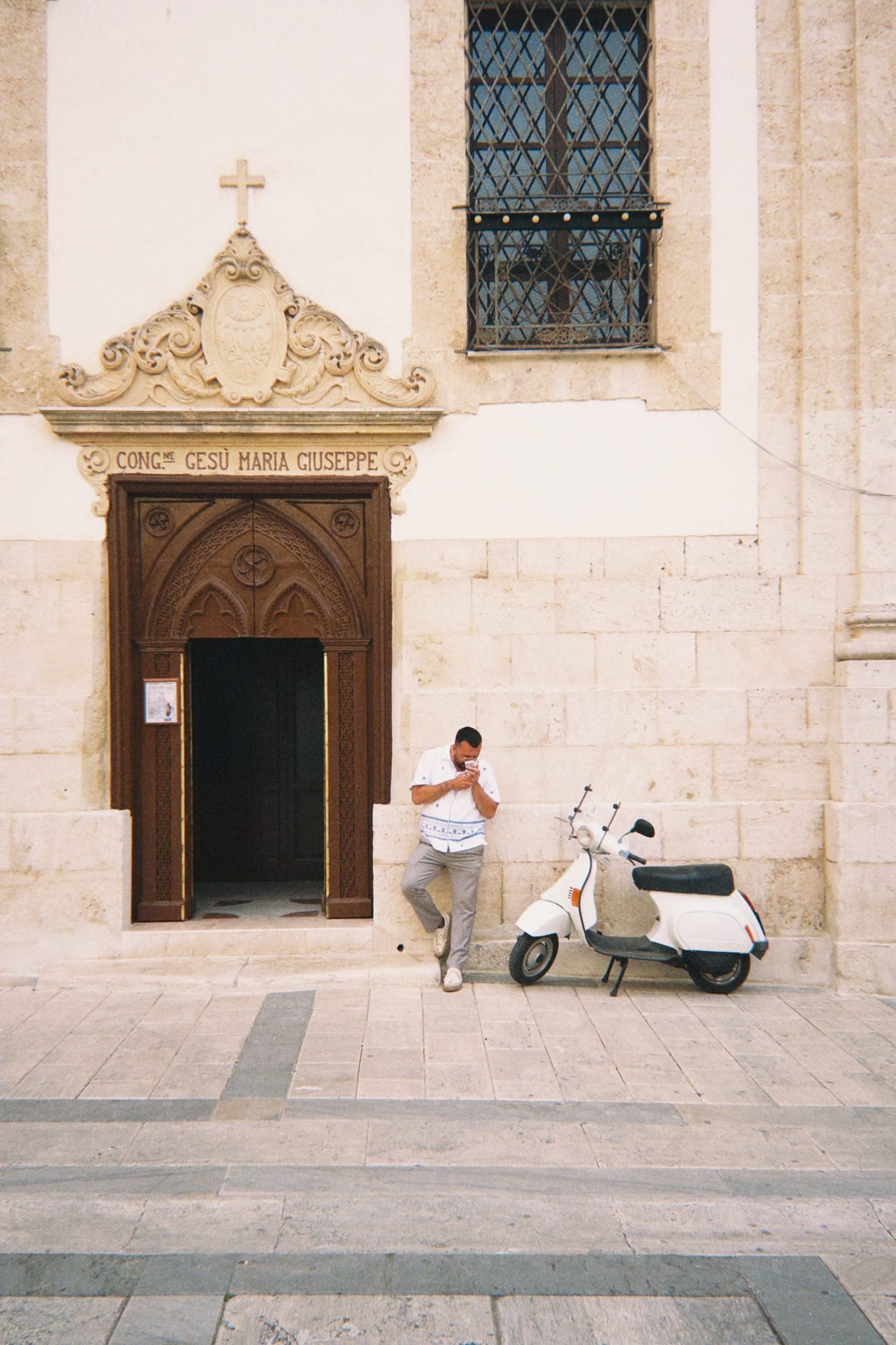 A man leans on a church wall in Alcamo beside a white Vespa, below the inscription “Cong.ne Gesù Maria Giuseppe” over the door