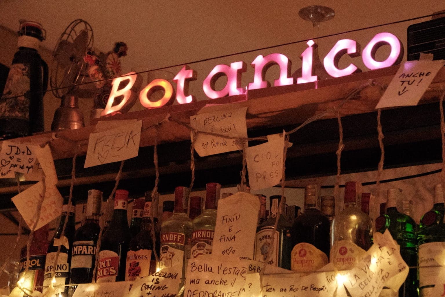 Pink neon sign reading “Botanico” above a shelf of bottles, handwritten notes on strings and fairy lights in front