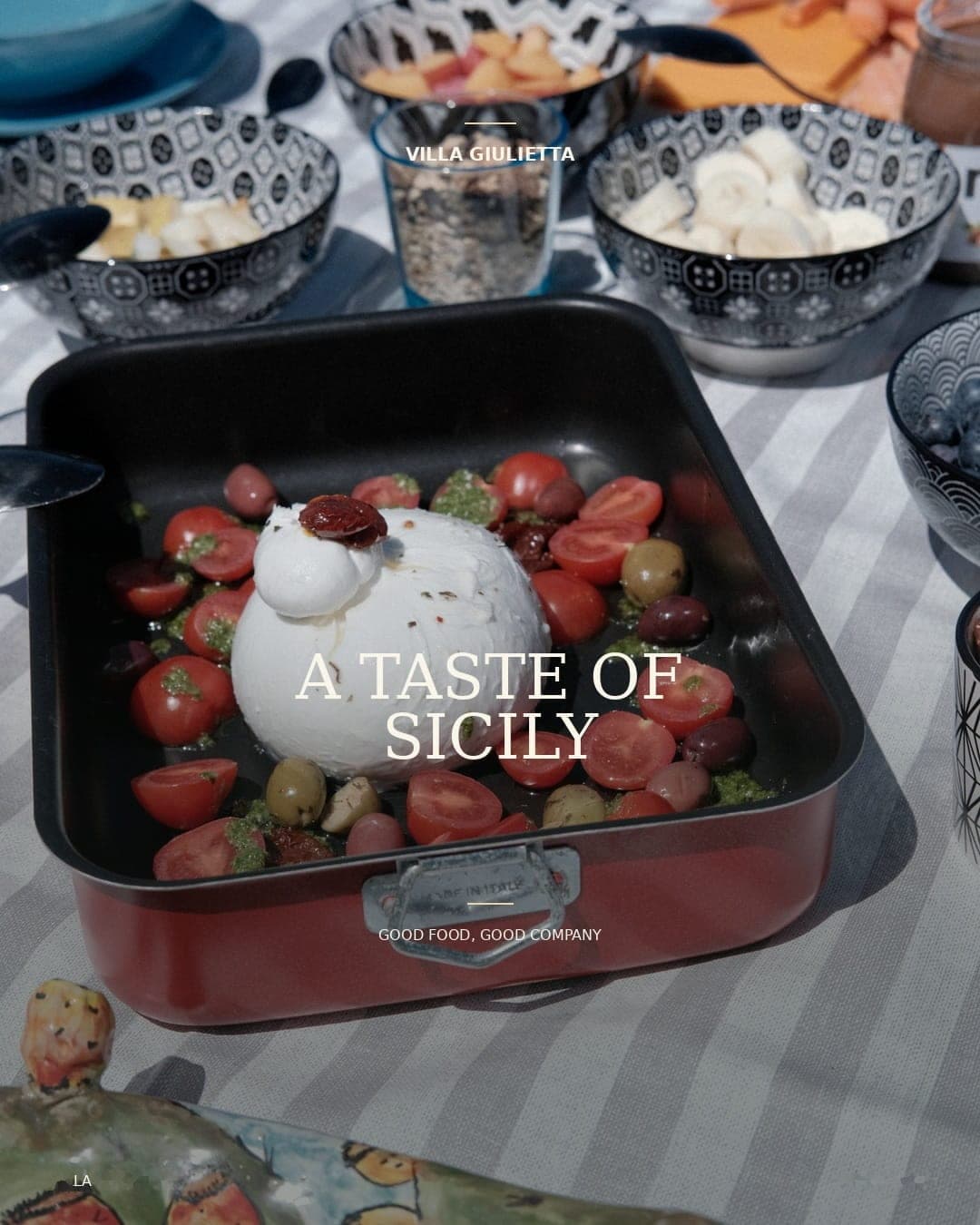 Burrata with cherry tomatoes and olives in a roasting tin among bowls of fruit, with the words “A taste of Sicily, good food, good company”