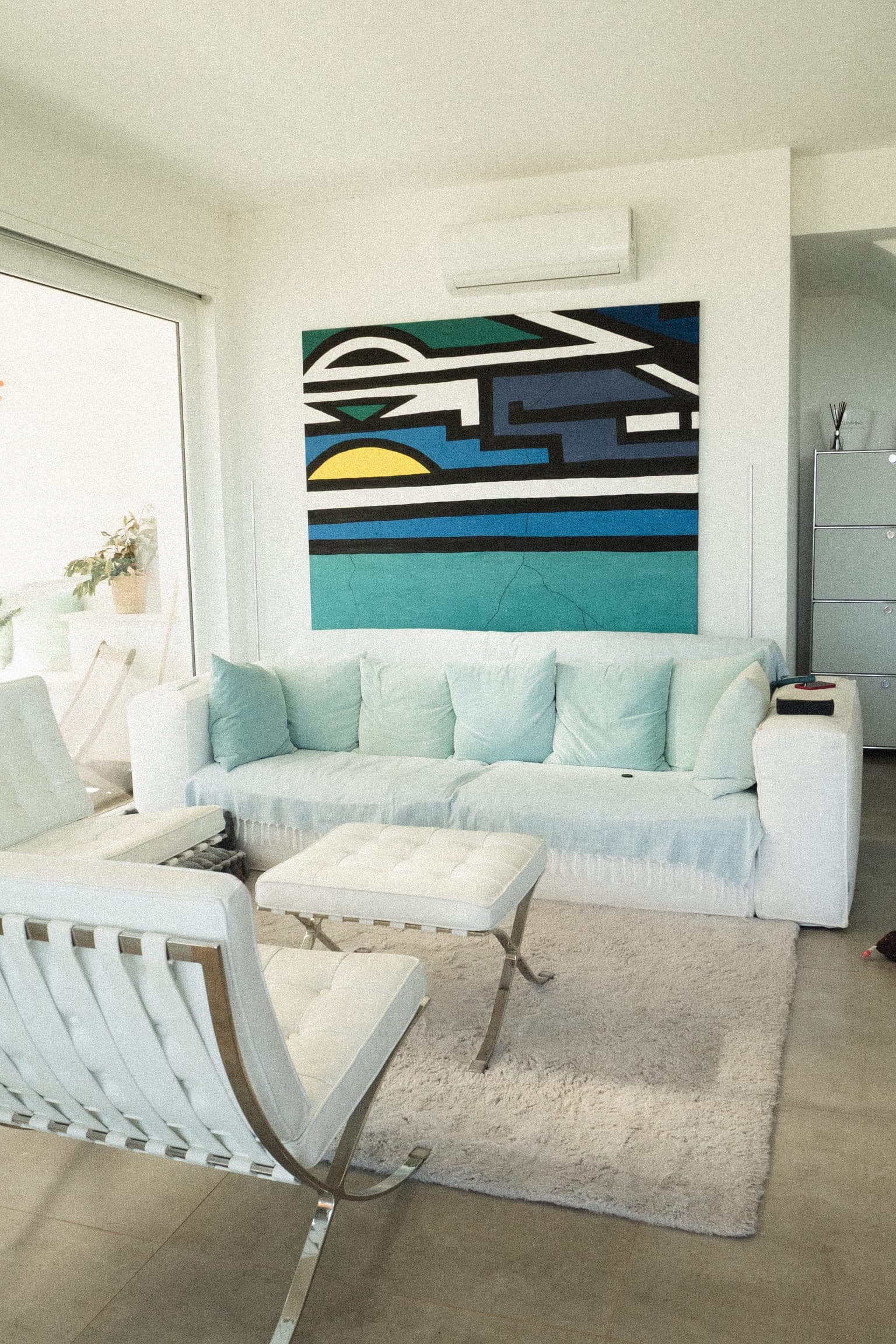 White sofa with pale green cushions below the large geometric wall painting, a white leather armchair and footstool on a rug in front