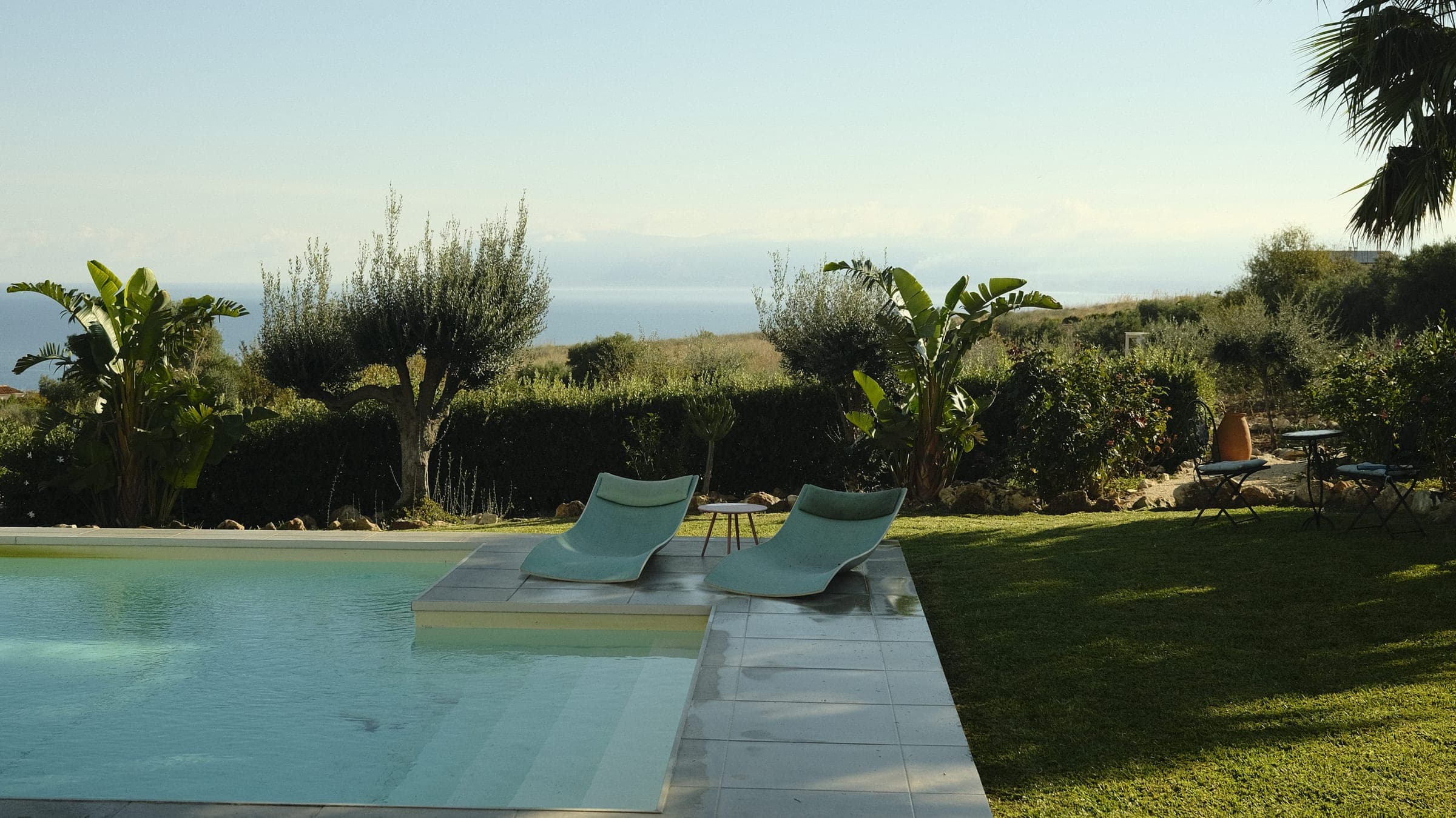 Two sun loungers at a corner of the pool, olive trees and a hedge behind them, the sea and the coast in the distance