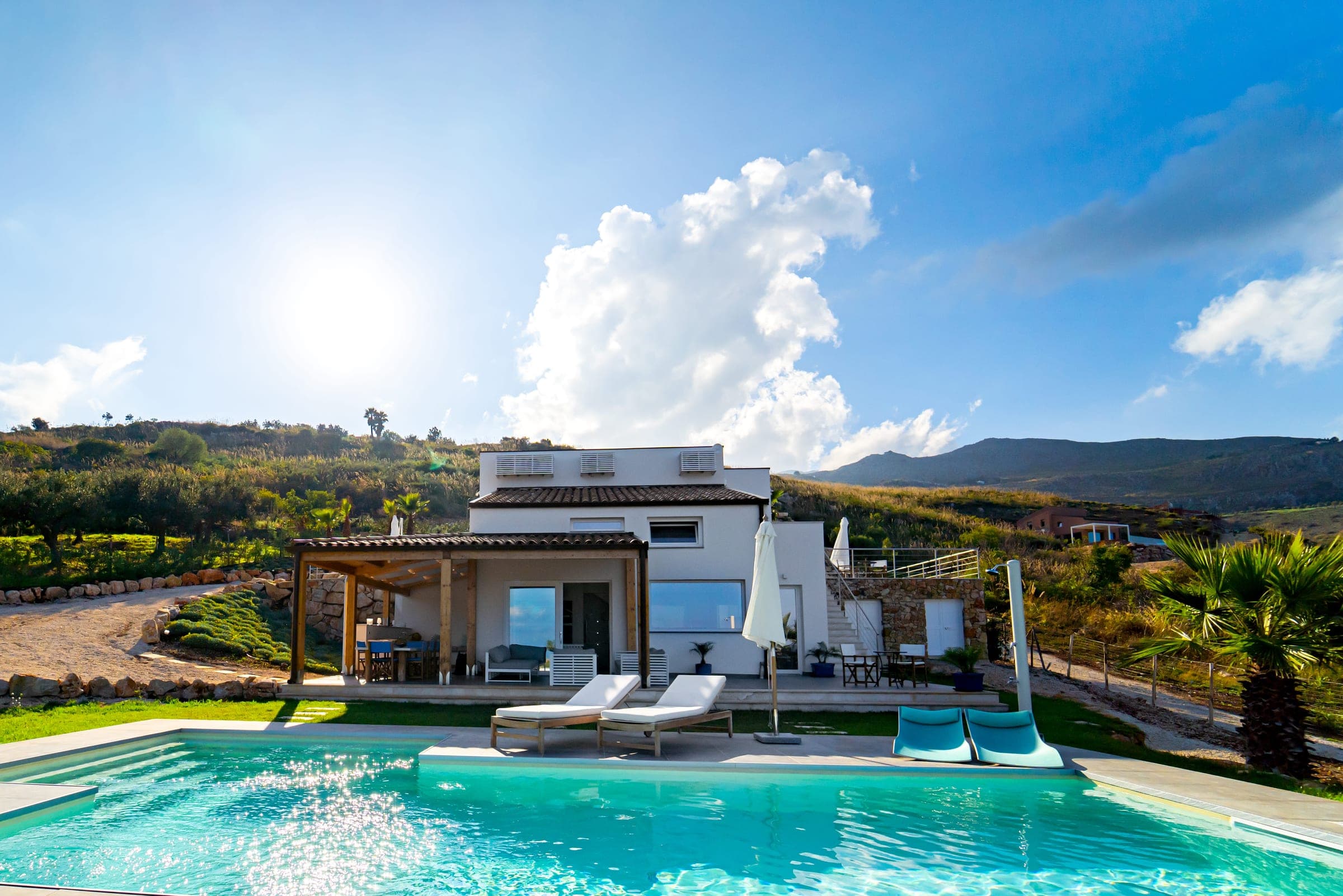 Pool and sun loungers in front of the white villa and its pergola, hills in the background