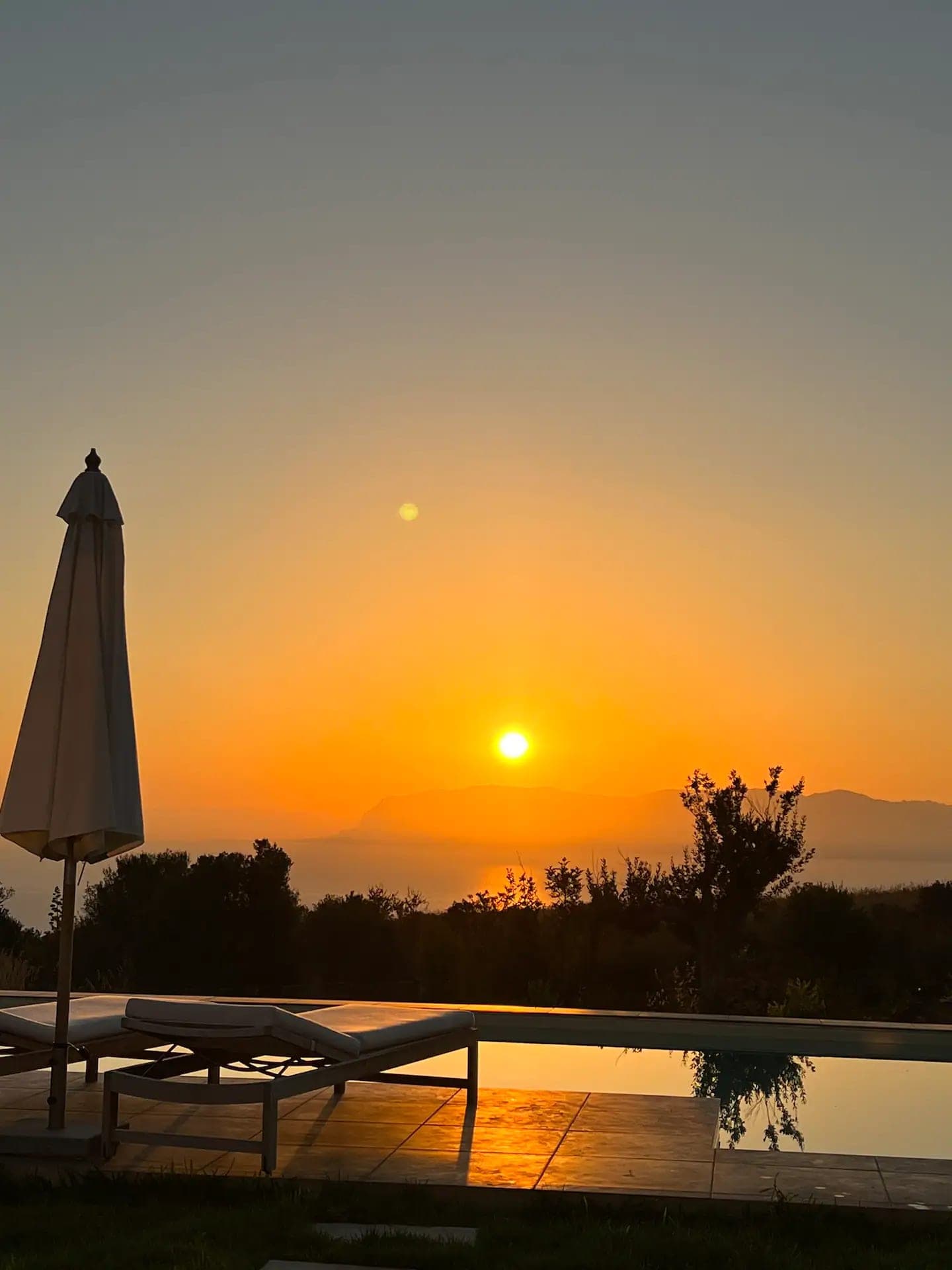 Sunset over the sea from the pool, with a closed parasol and a sun lounger in the foreground