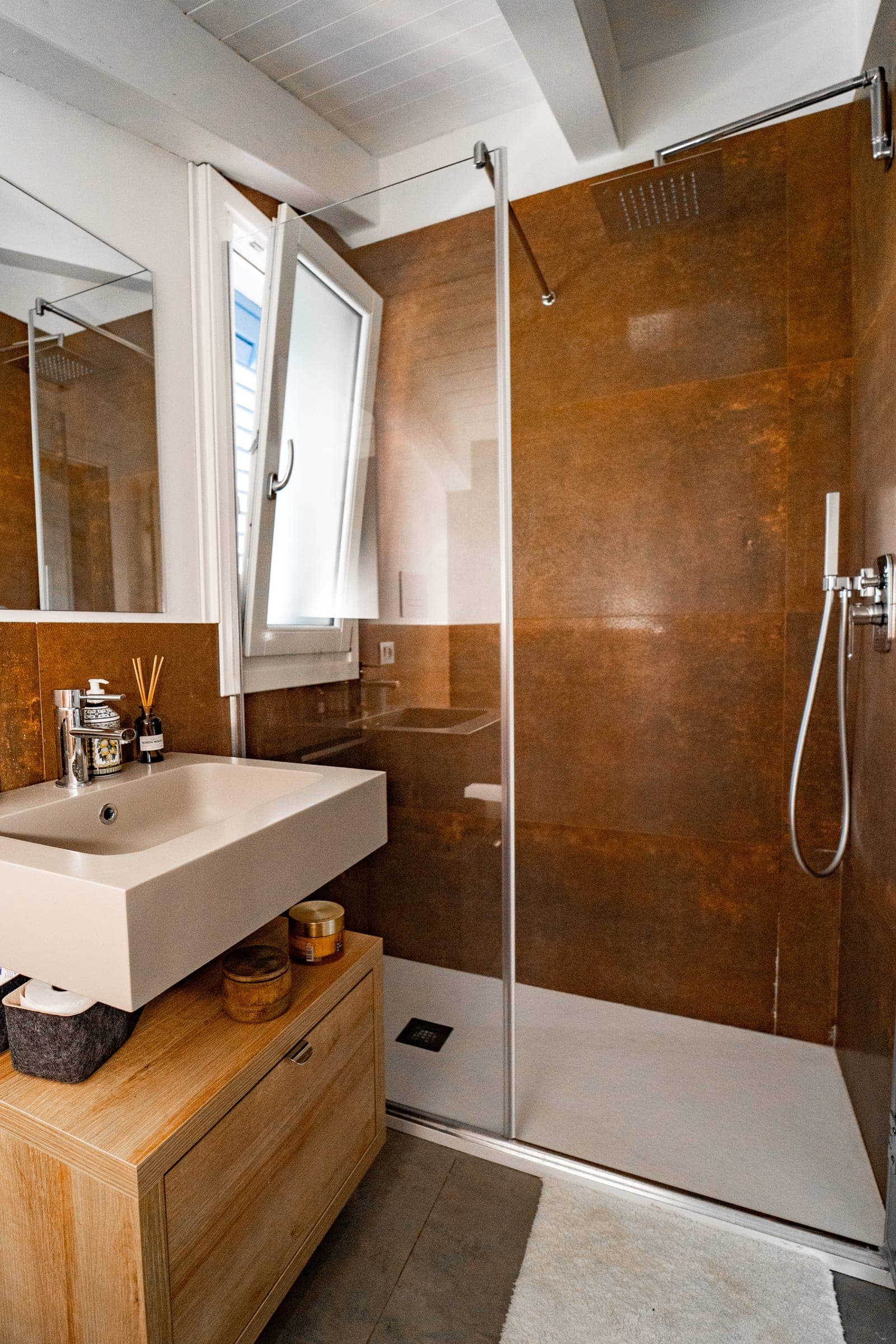 Shower with rust-coloured tiles behind a glass screen, next to a white washbasin on a wooden unit